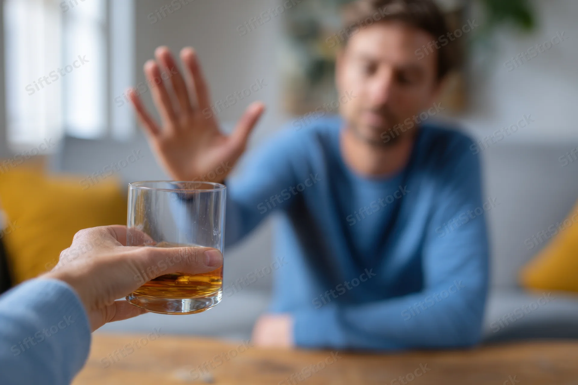 Man in blue long-sleeve shirt refusing a glass of whiskey at a wooden table in a living room