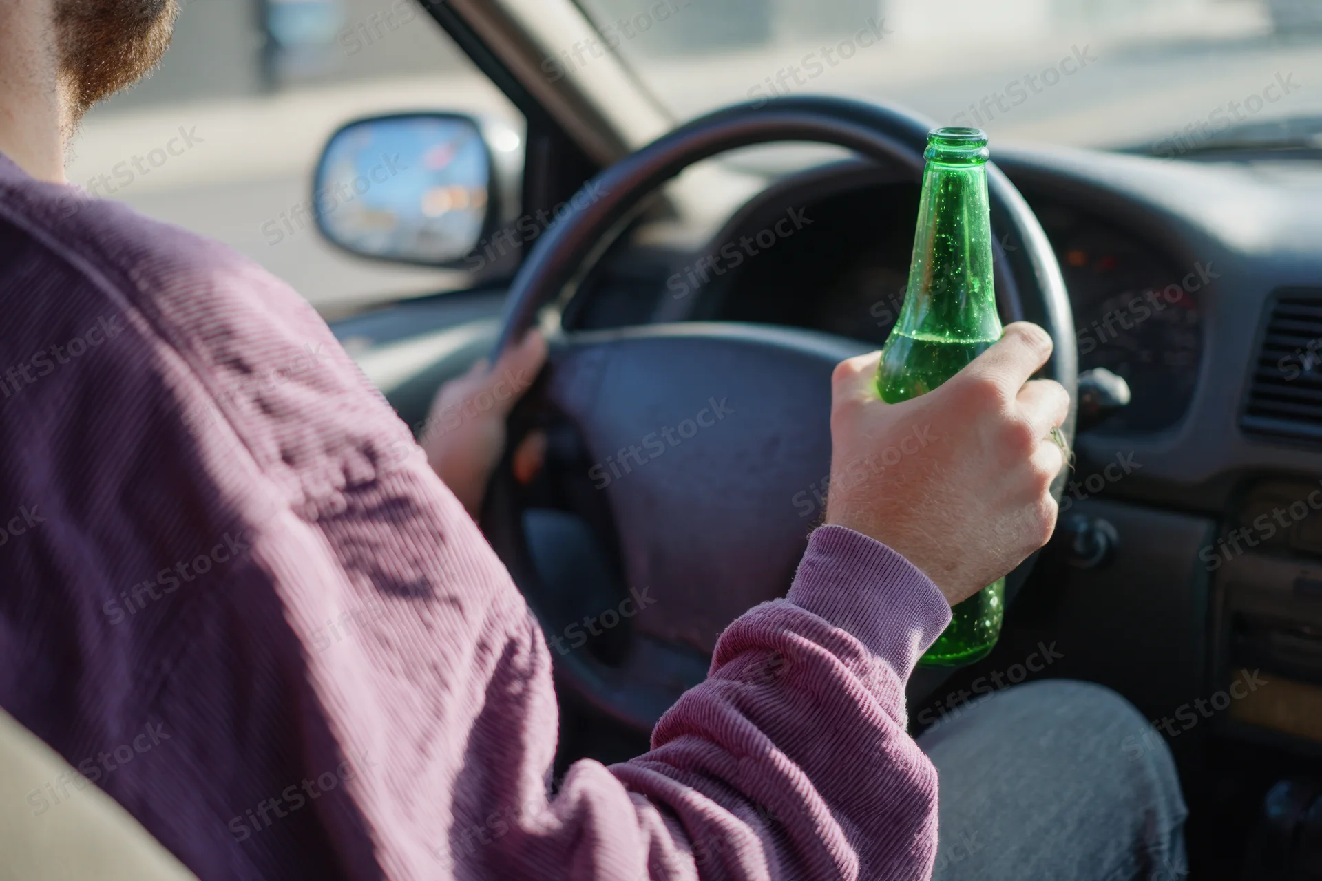 Man driving a car while holding a green beer bottle in his hand, viewed from over the shoulder inside the vehicle