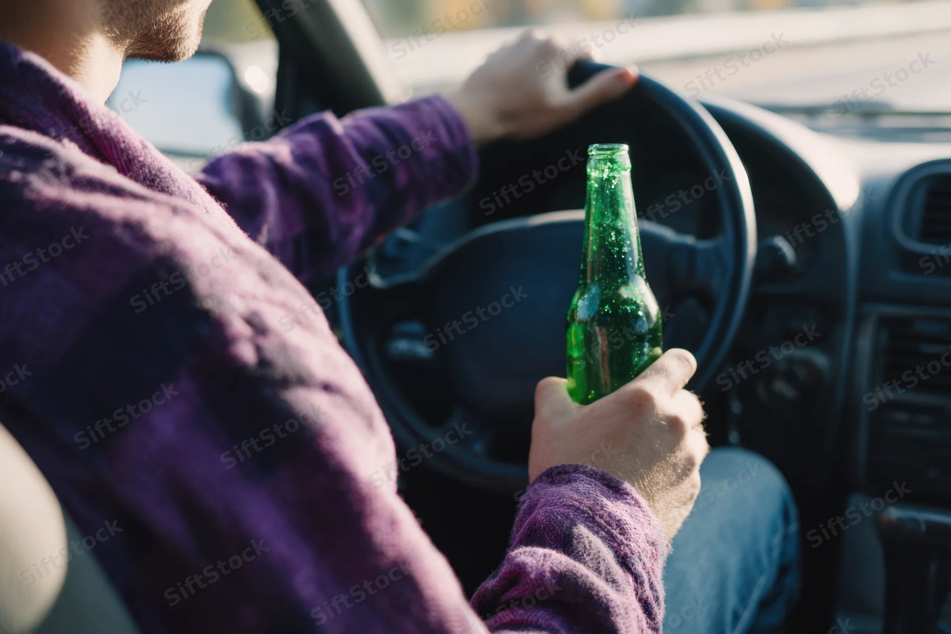 Man driving a car while holding a green beer bottle in his hand