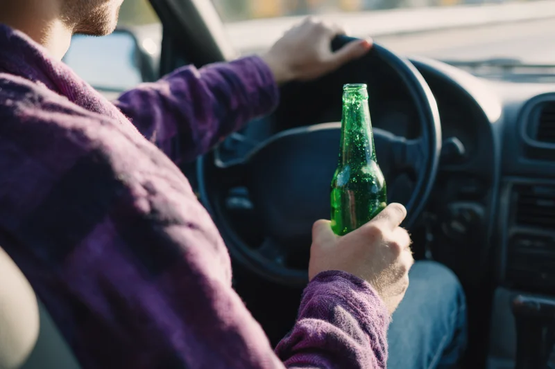 Man driving a car while holding a green beer bottle in his hand