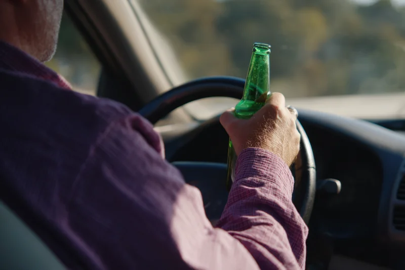 Man driving car holding green glass beer bottle in hand on steering wheel