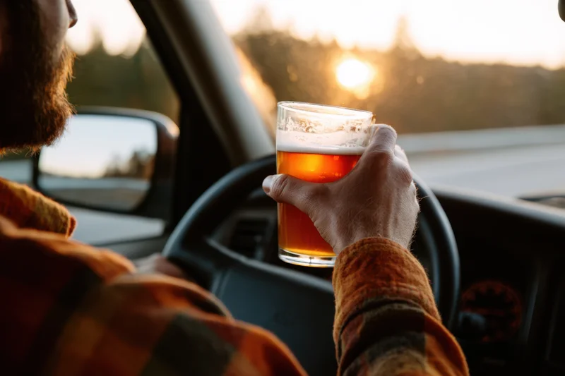 Man holding a glass of beer while driving a car at sunset