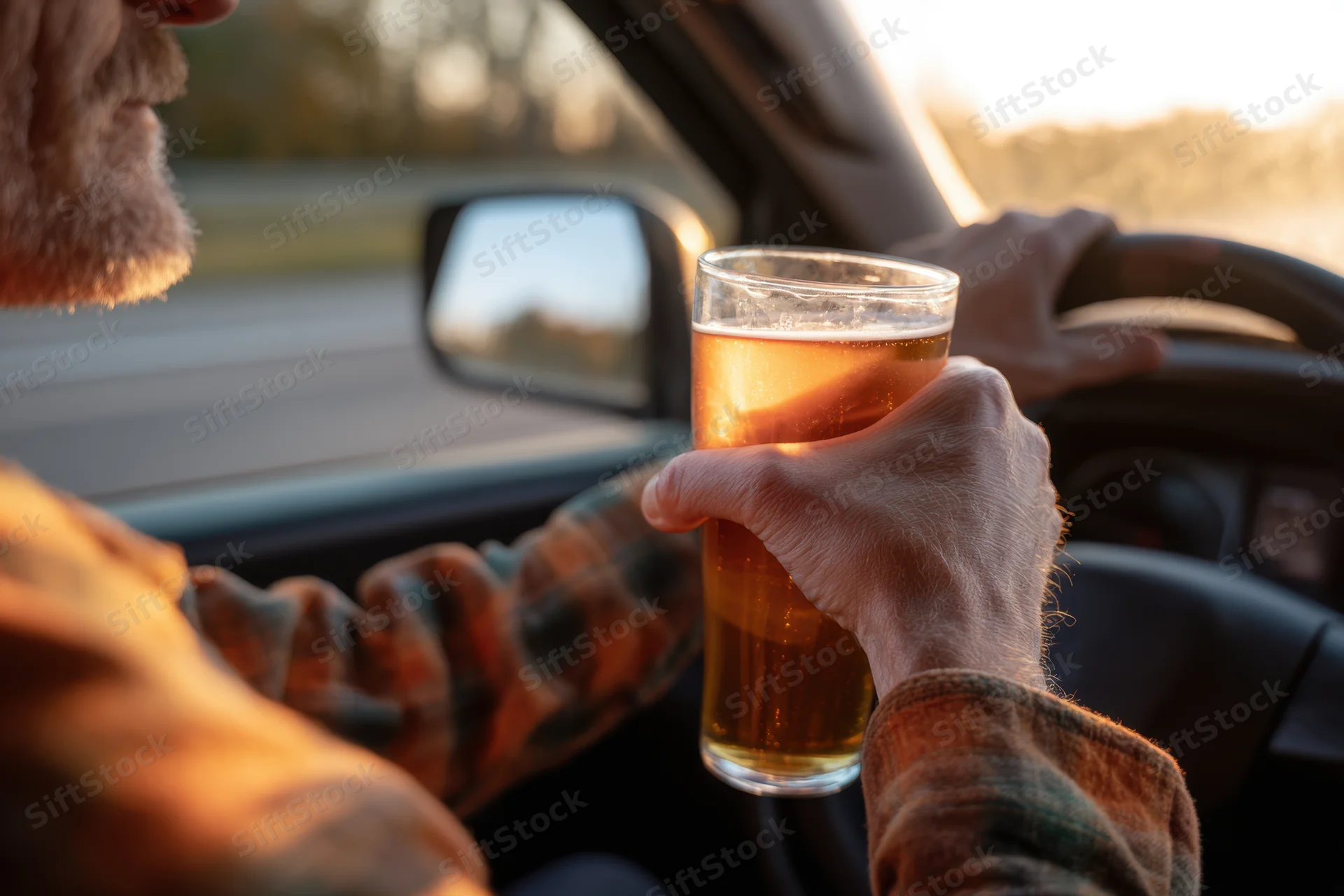 Man holding a glass of beer while driving a car at sunset