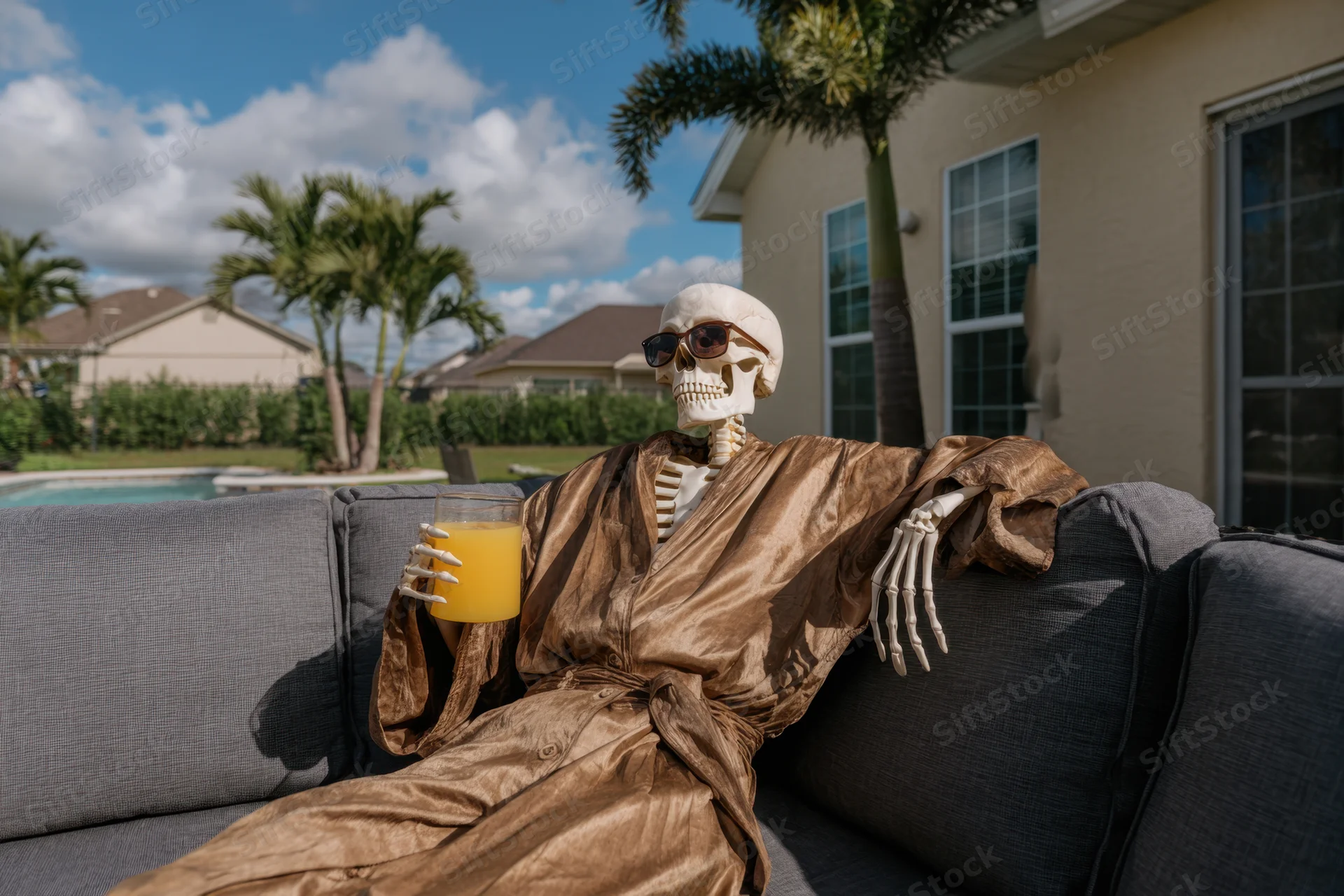 Skeleton wearing sunglasses and a robe lounging on a sofa in a backyard with a pool and palm trees