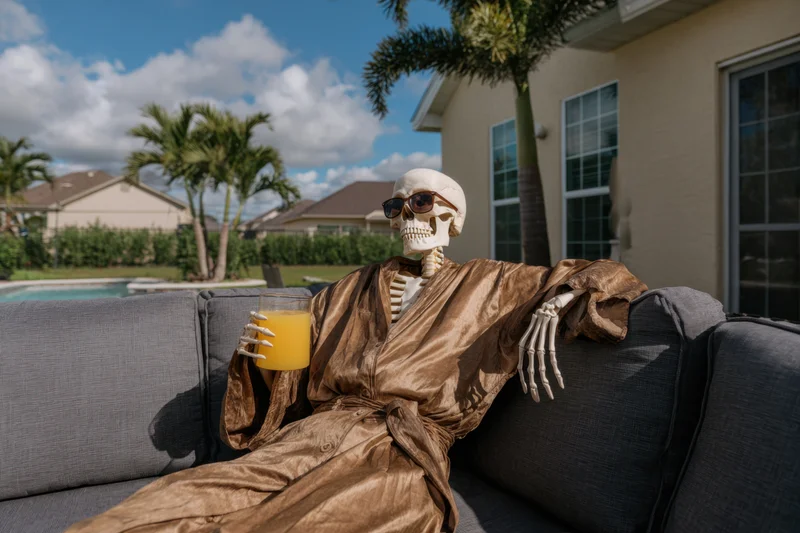 Skeleton wearing sunglasses and a robe lounging on a sofa in a backyard with a pool and palm trees