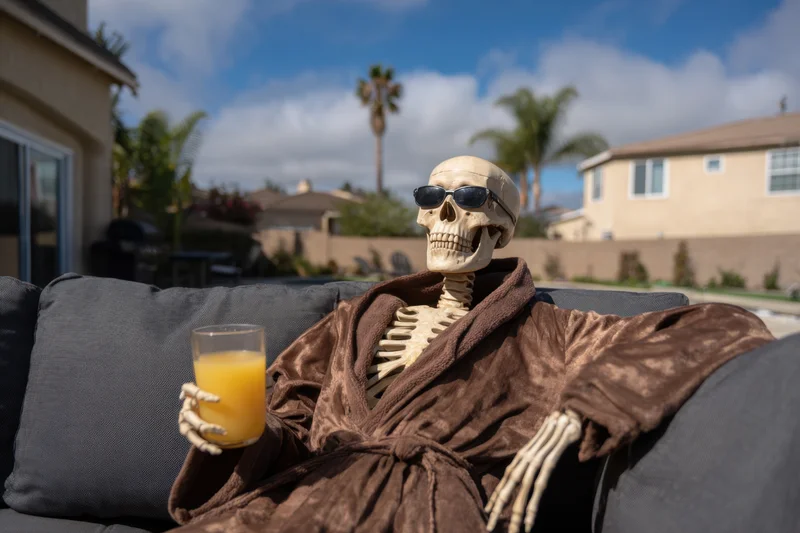 Skeleton wearing sunglasses and a robe, holding a glass of orange juice, lounging on a dark outdoor couch in a suburban backyard