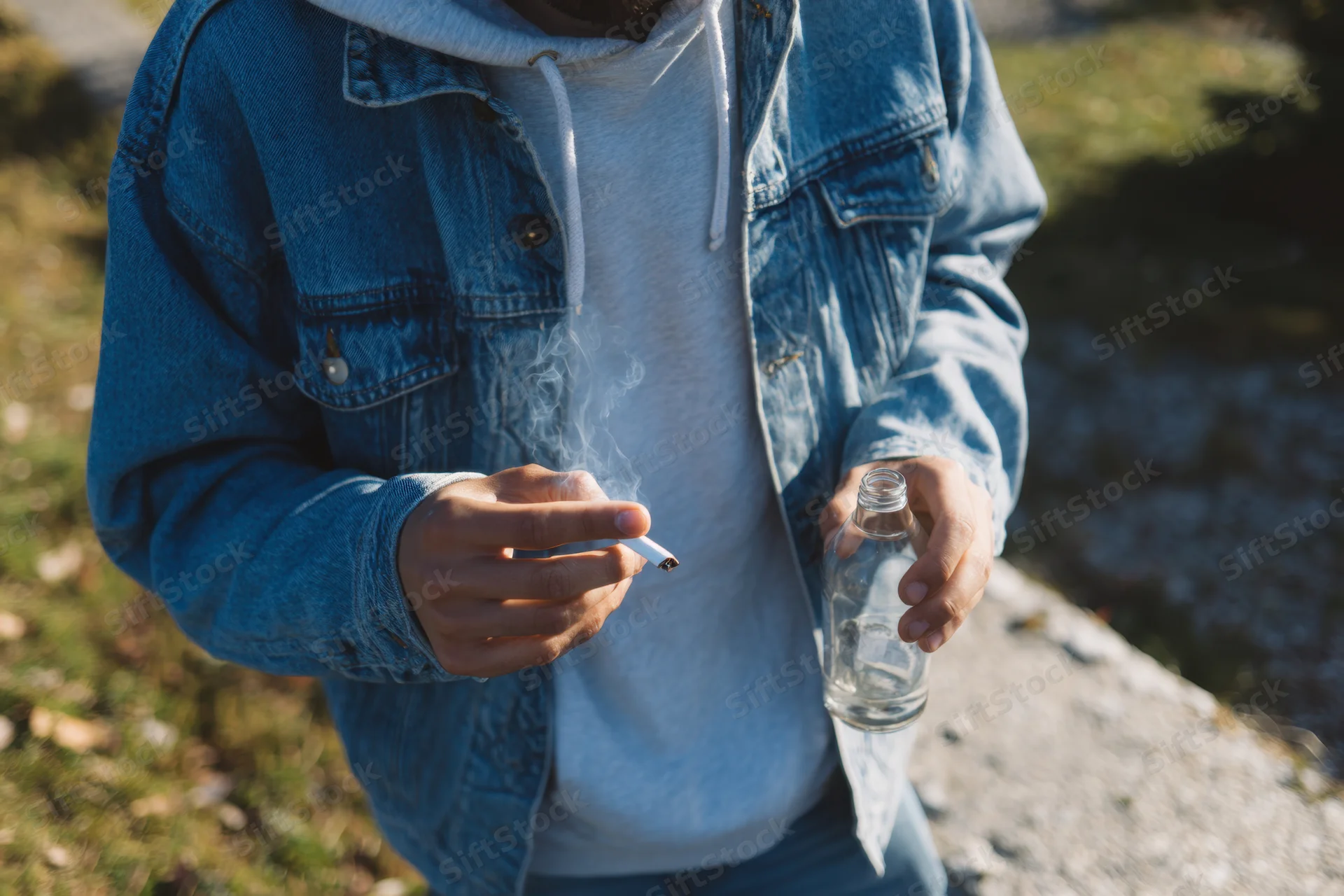 Young man smoking a cigarette and holding an empty glass bottle outdoors