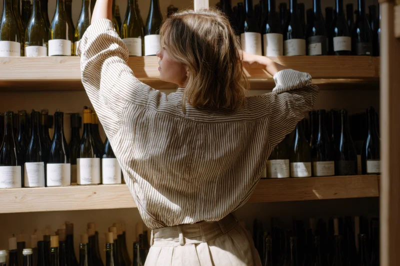Woman reaching for a bottle on a wooden shelf in a wine shop