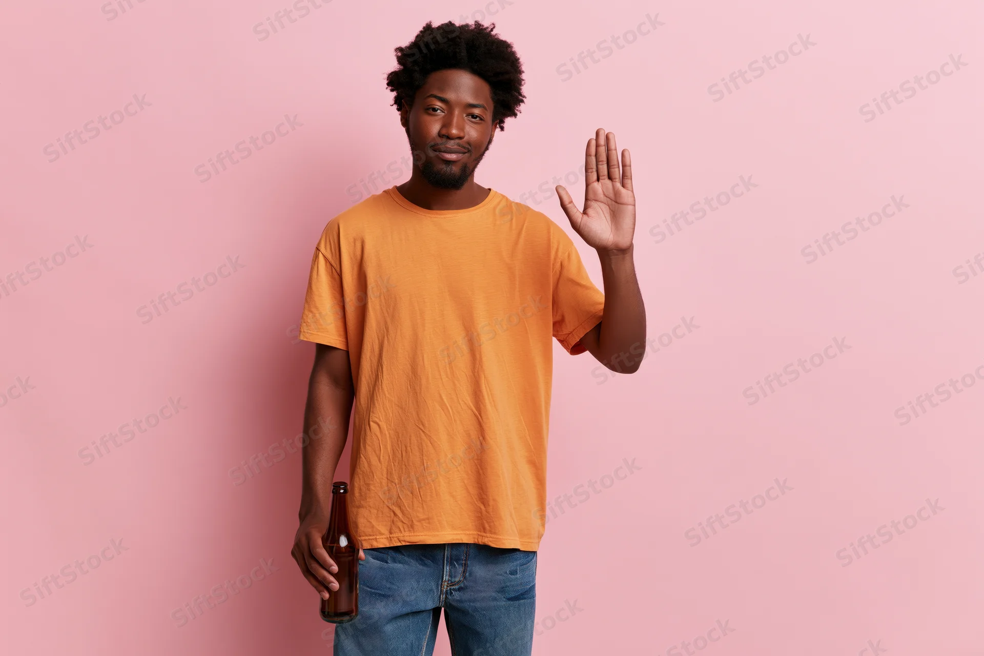 Young black man in orange tshirt and jeans waving while holding a beer bottle against pink background