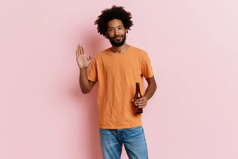 Young man in orange tshirt and jeans waving while holding a beer bottle against pink background