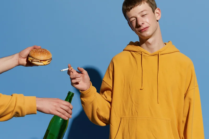 Young man in yellow hoodie holding cigarette with burger and beer offered to him in studio