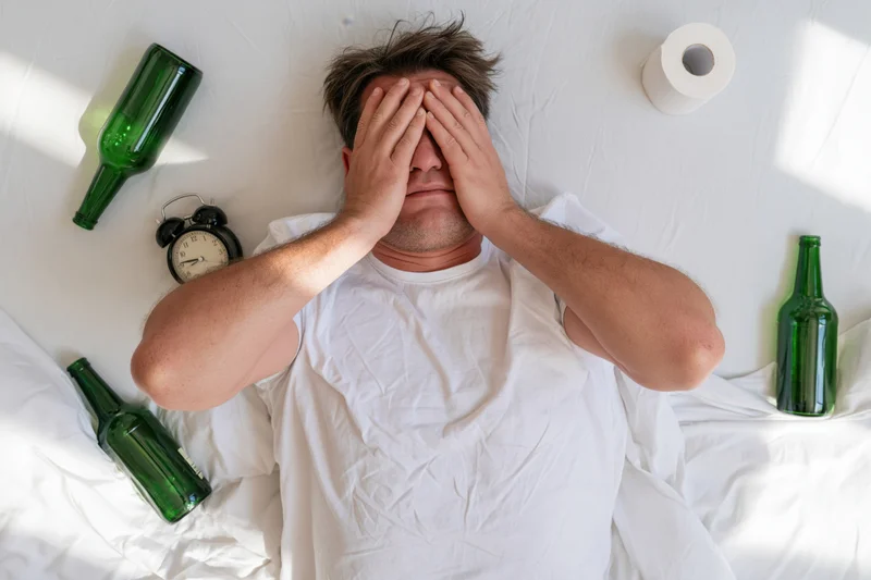 Man lying on white bed covering face with hands surrounded by empty green bottles and alarm clock