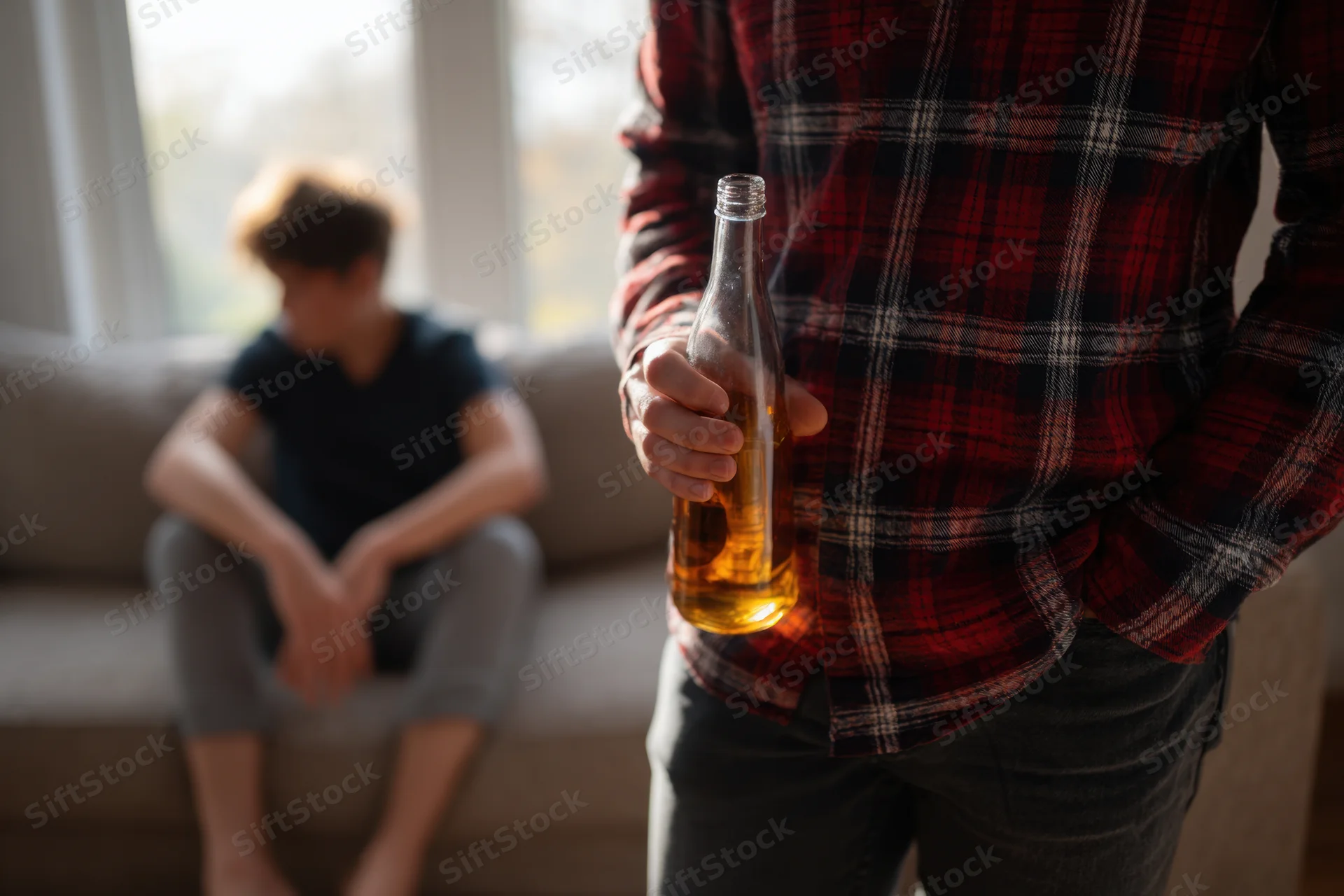 Man holding a beer bottle in a living room with a blurred person sitting on a couch in the background