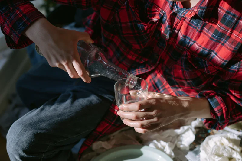 Person pouring liquid from glass bottle into glass cup while seated indoors