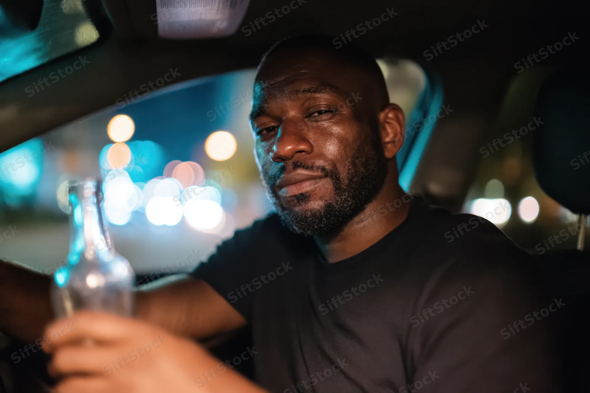 Black man holding glass bottle in car at night with city lights blurred in background