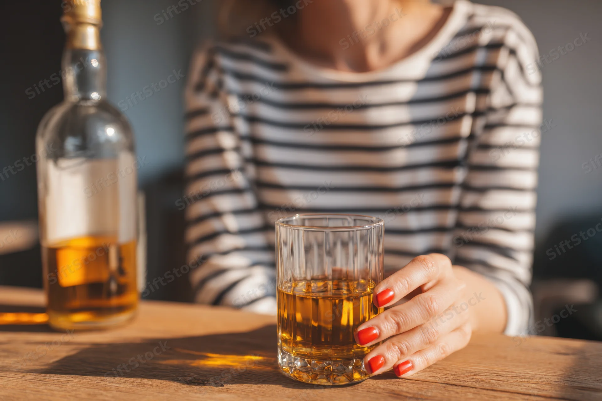 Woman holding a glass of whiskey on a wooden table with a bottle nearby