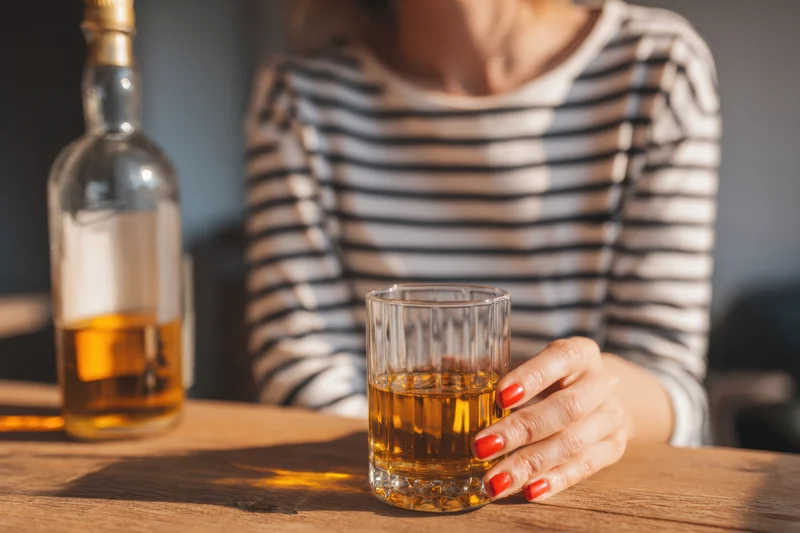 Woman holding a glass of whiskey on a wooden table with a bottle nearby
