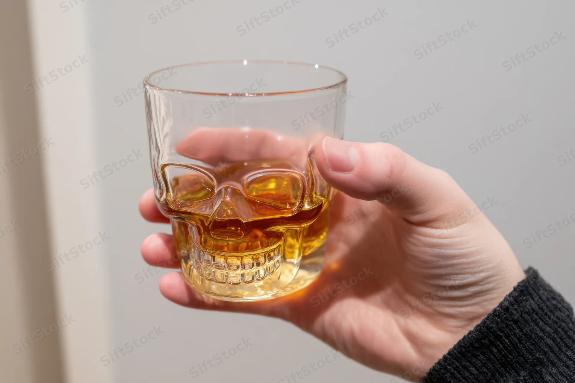 Hand holding a skull-shaped glass filled with amber whiskey against a plain background