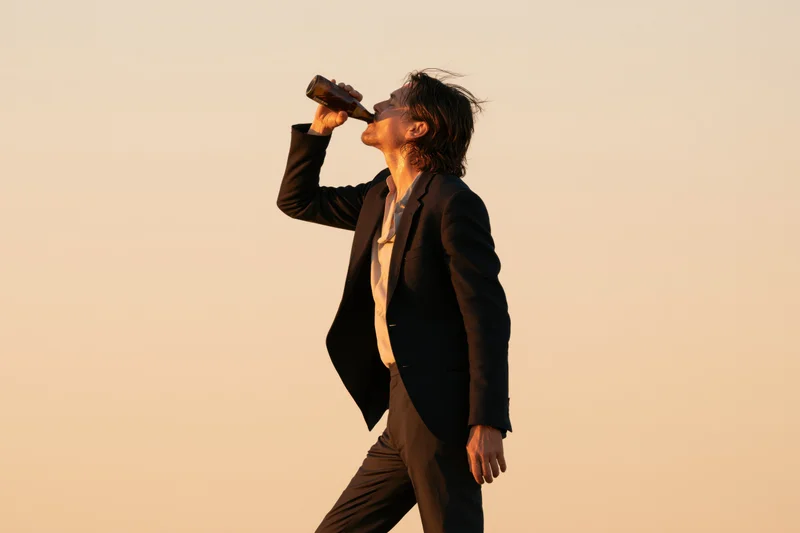 Man in dark blazer drinking beer outdoors at sunset