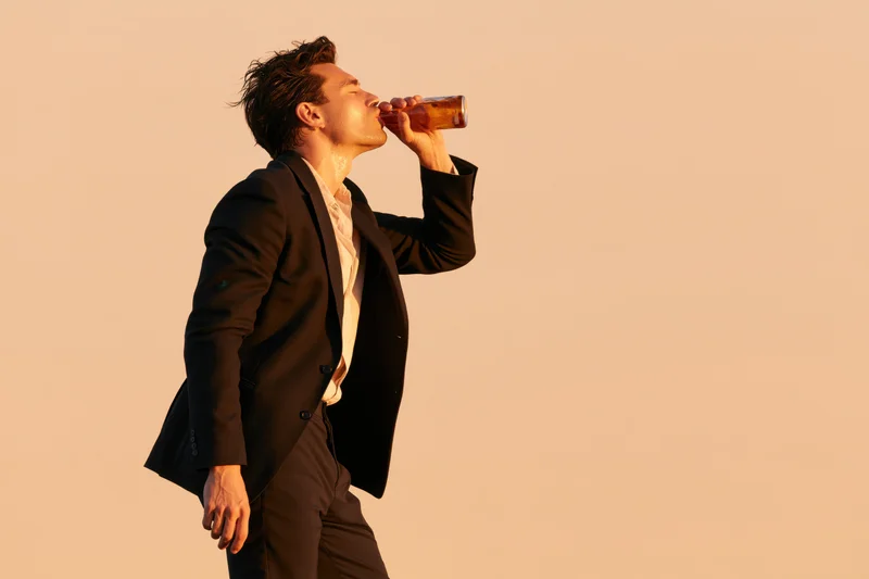 Man in dark blazer drinking beer from bottle in golden hour light with copyspace to the right