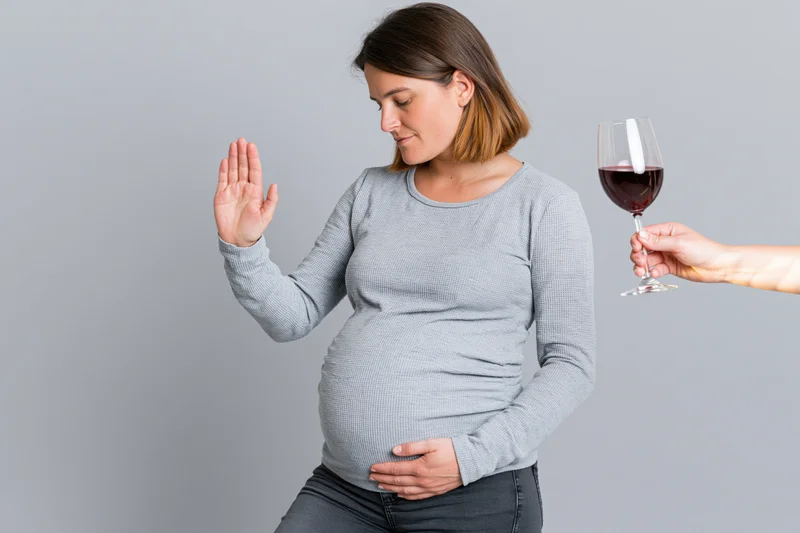 Pregnant woman refusing a glass of red wine while holding her belly against a gray background