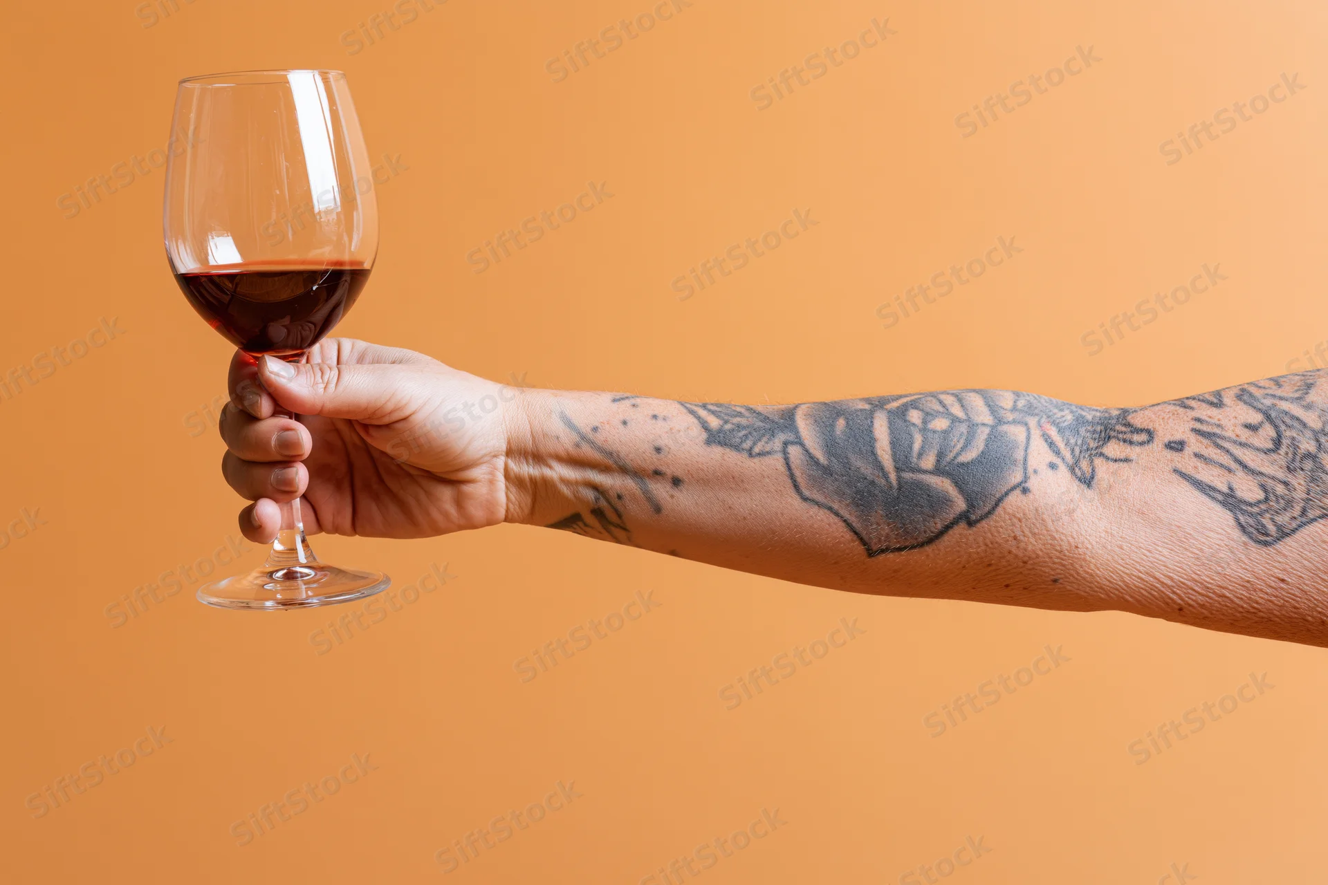 Tattooed arm holding a glass of red wine against orange background