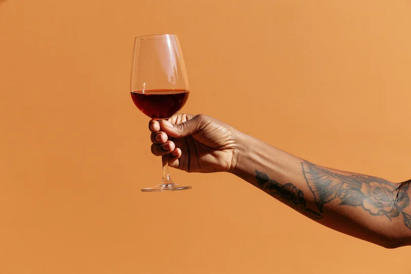 Tattooed hand holding a glass of red wine against an orange background