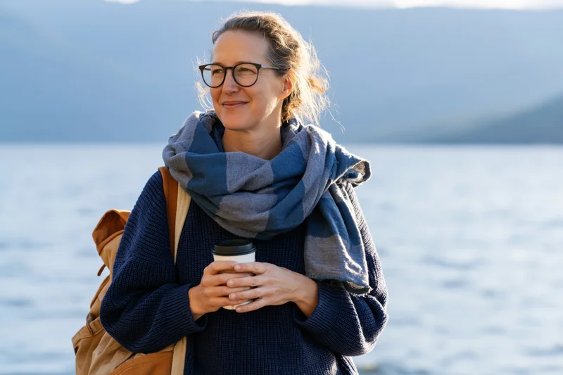 Woman smiling while holding coffee cup by lake with mountains in background