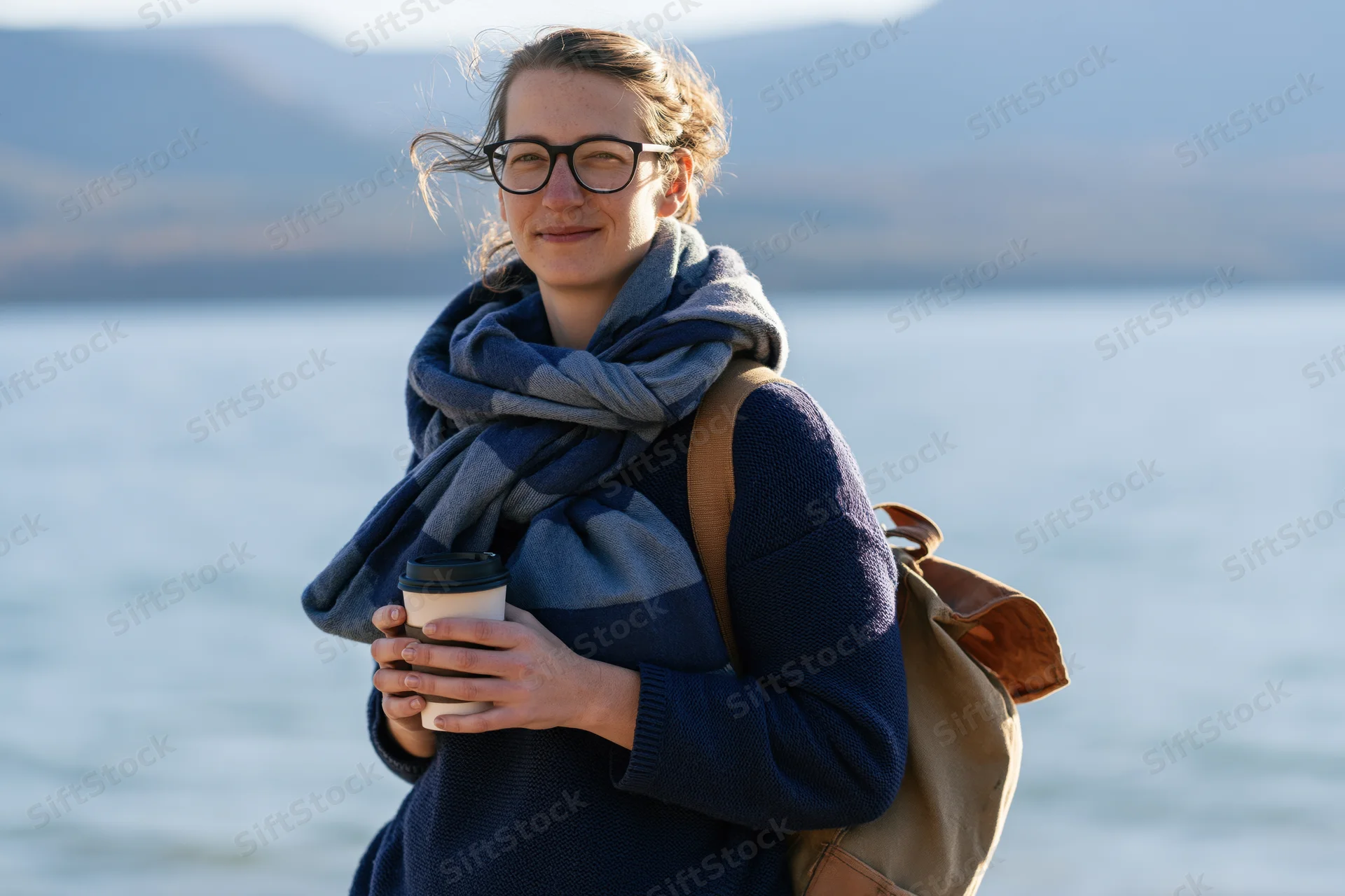 Woman holding coffee mug by lake with backpack and scarf, smiling in wind
