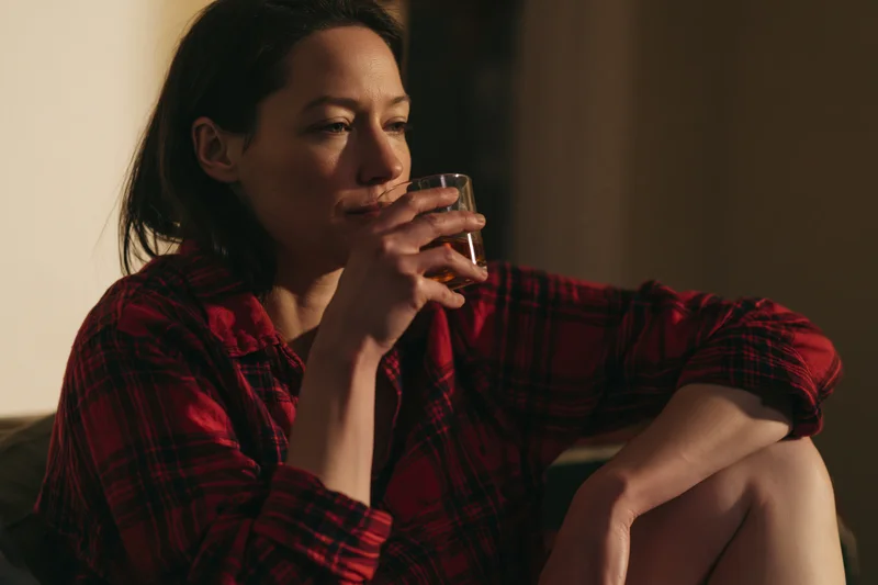 Woman drinking whiskey from a glass while seated on a couch in a dimly lit room