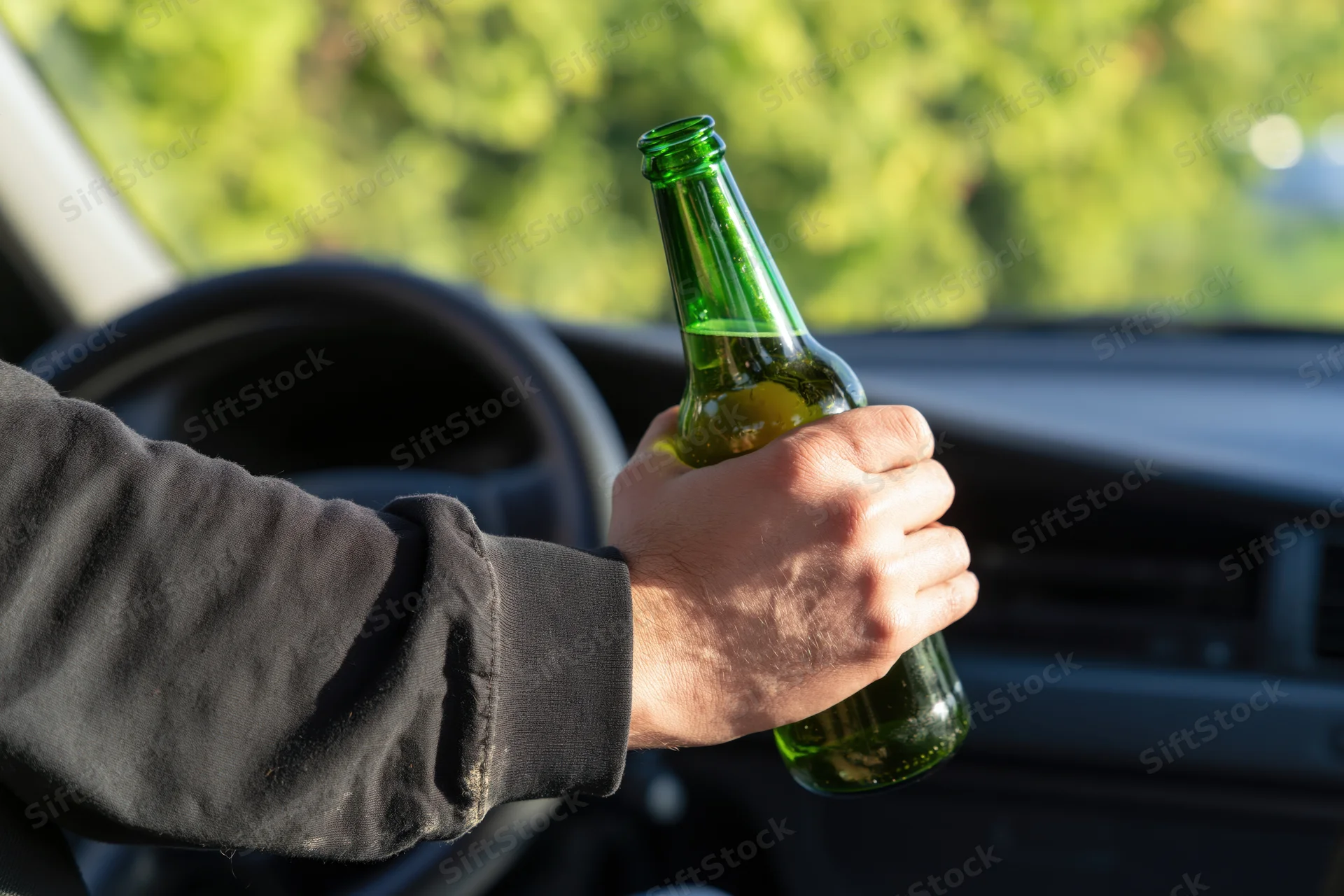 Hand holding a green glass beer bottle inside a car near the steering wheel