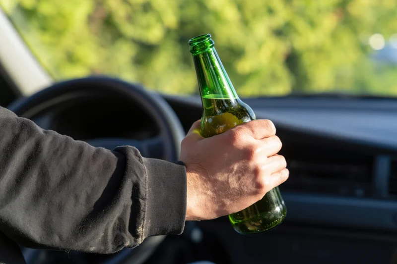 Hand holding a green glass beer bottle inside a car near the steering wheel