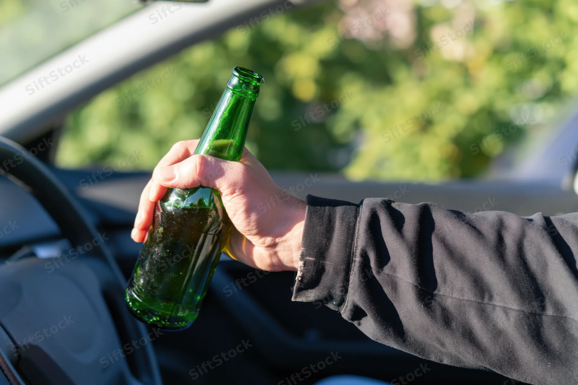 Hand holding green glass beer bottle inside car near steering wheel