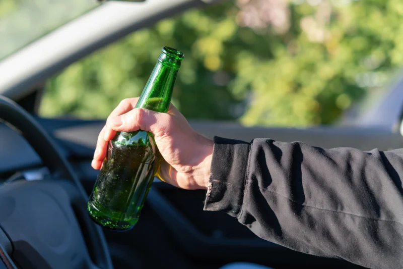 Hand holding green glass beer bottle inside car near steering wheel