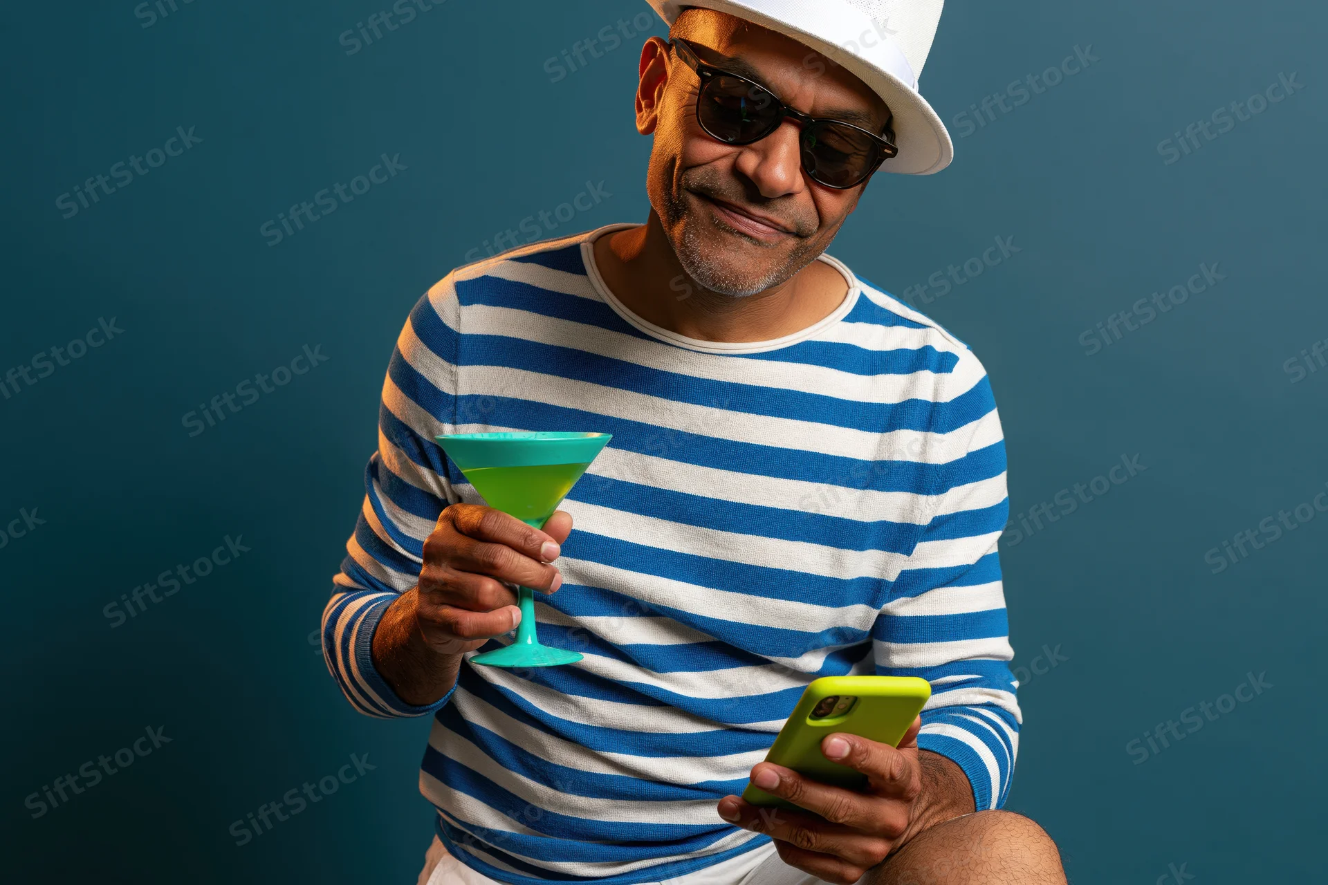 Man in striped shirt and hat holding cocktail and smartphone, smiling against blue background