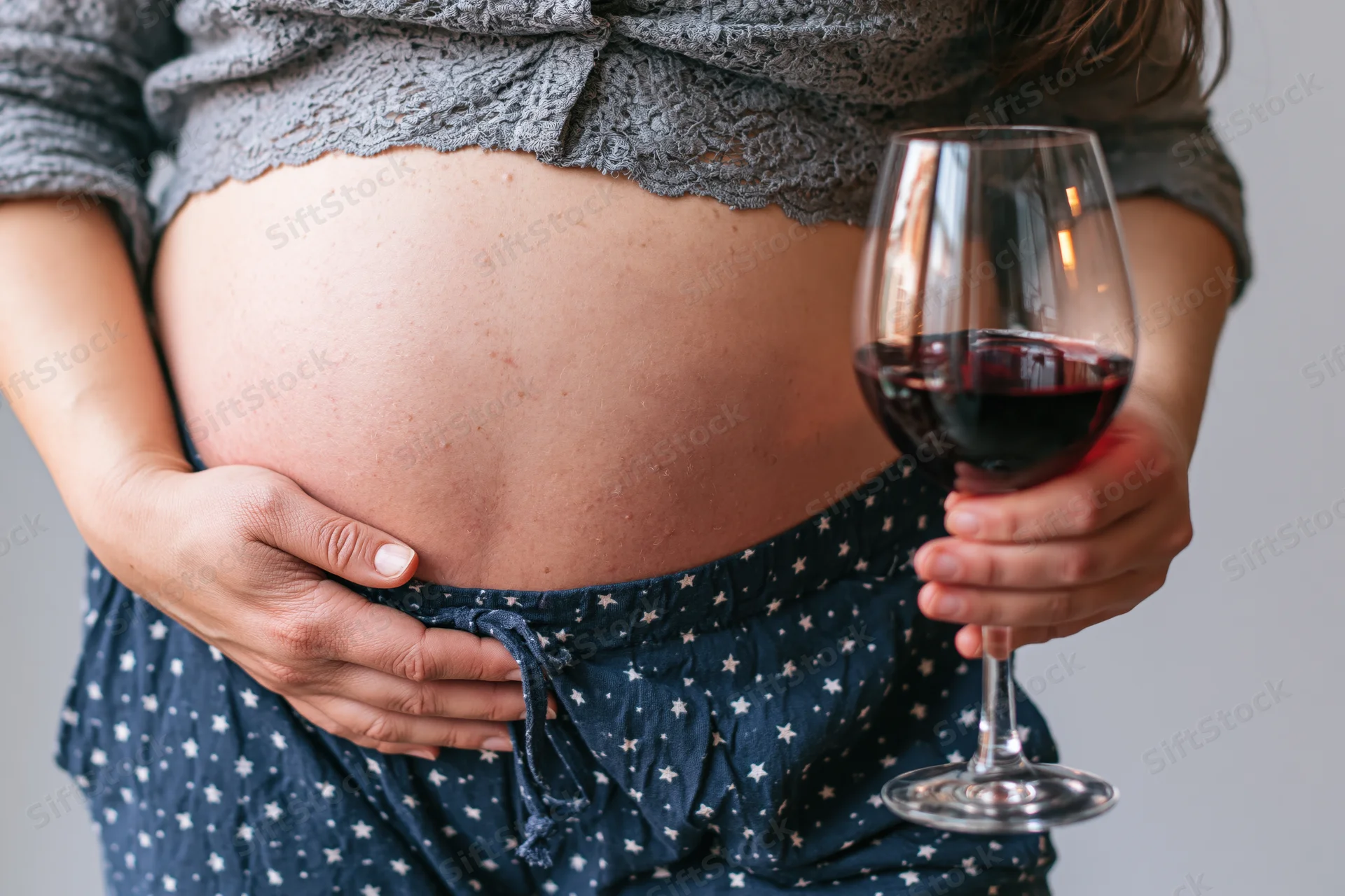Pregnant woman holding a glass of red wine while cradling her belly indoors