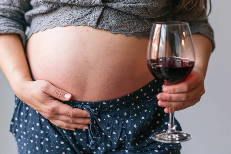 Pregnant woman holding a glass of red wine while cradling her belly indoors