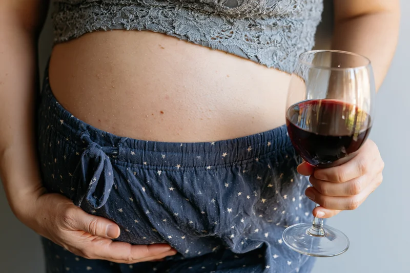Pregnant woman holding a glass of red wine while cradling her belly indoors