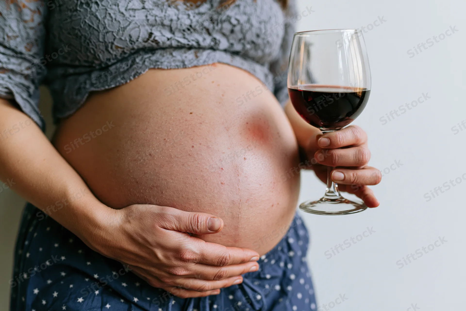 Pregnant woman holding a glass of red wine while cradling her belly indoors