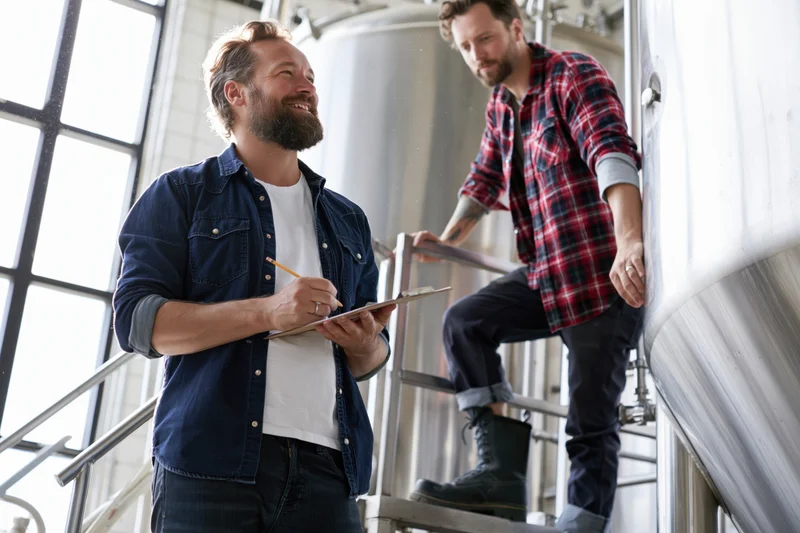 Two men inspecting stainless steel tanks in a brewery with one holding a clipboard and pencil