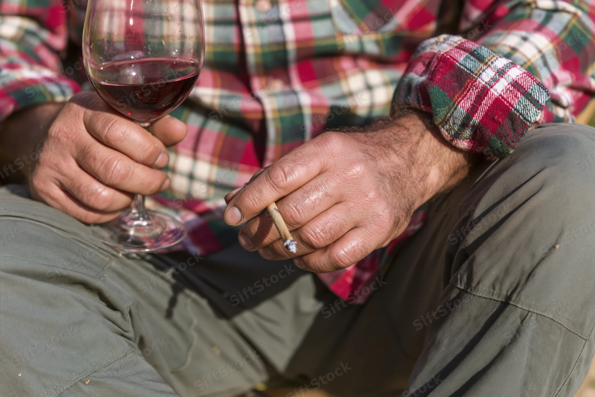 Man holding a glass of red wine and a lit cigarette while seated outdoors in a plaid shirt