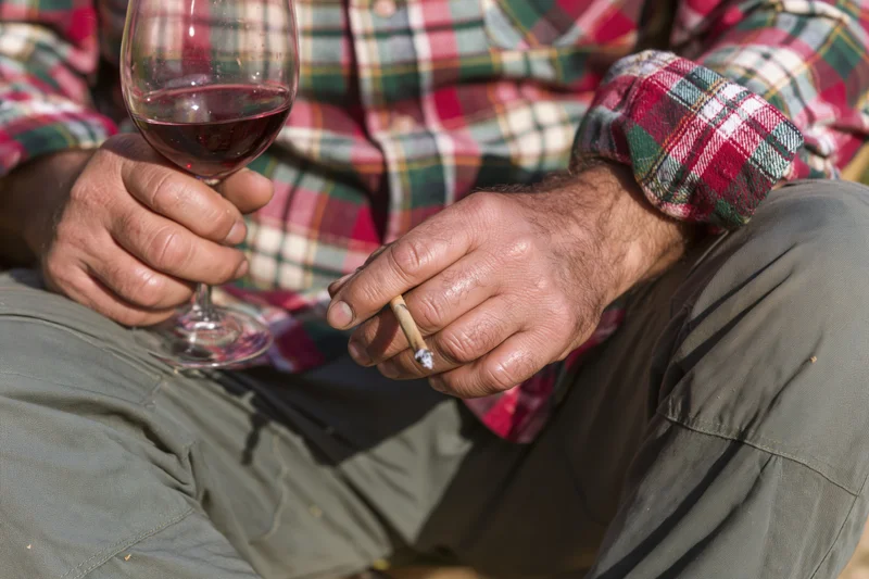 Man holding a glass of red wine and a lit cigarette while seated outdoors in a plaid shirt