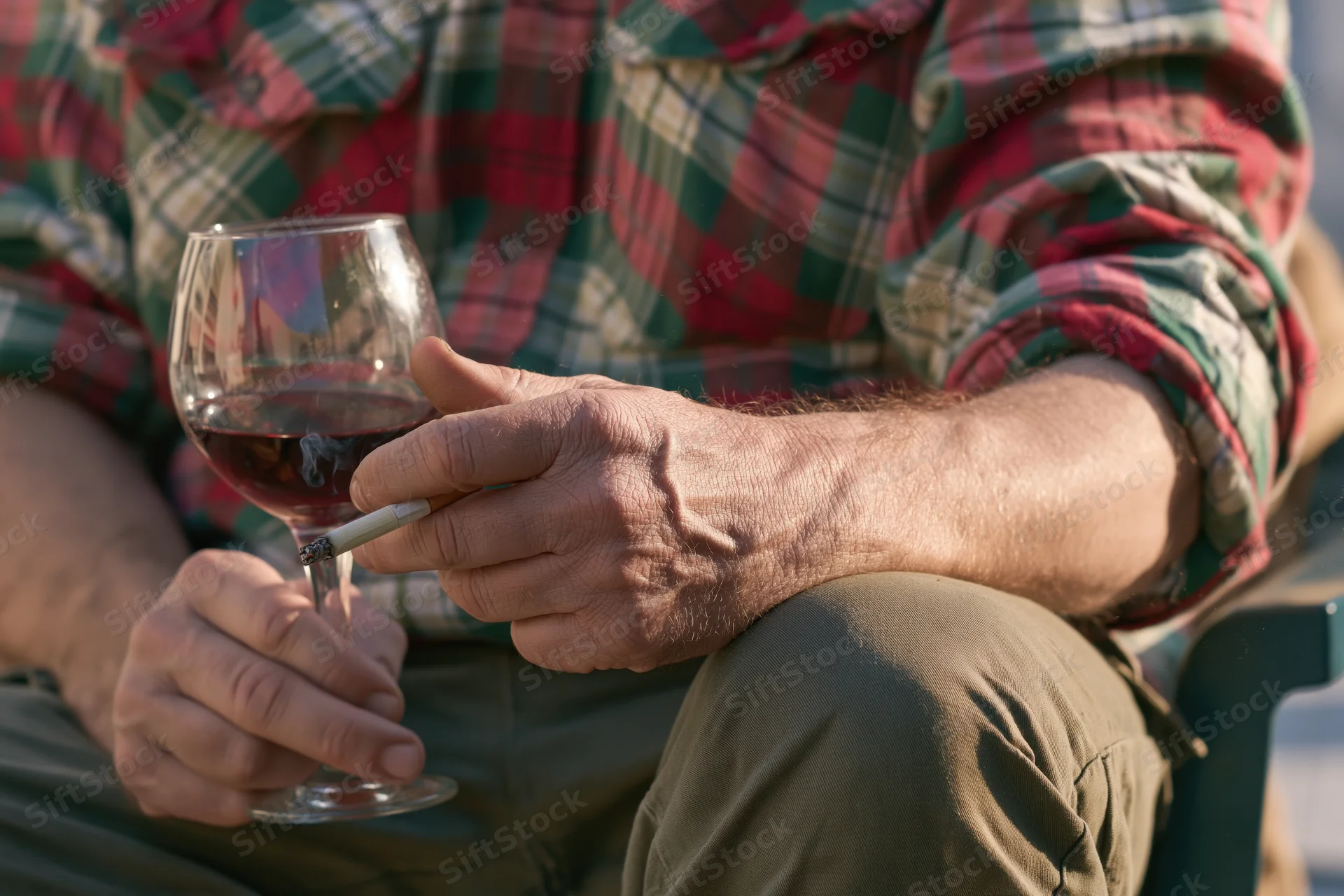 Man holding a glass of red wine and a cigarette while seated outdoors