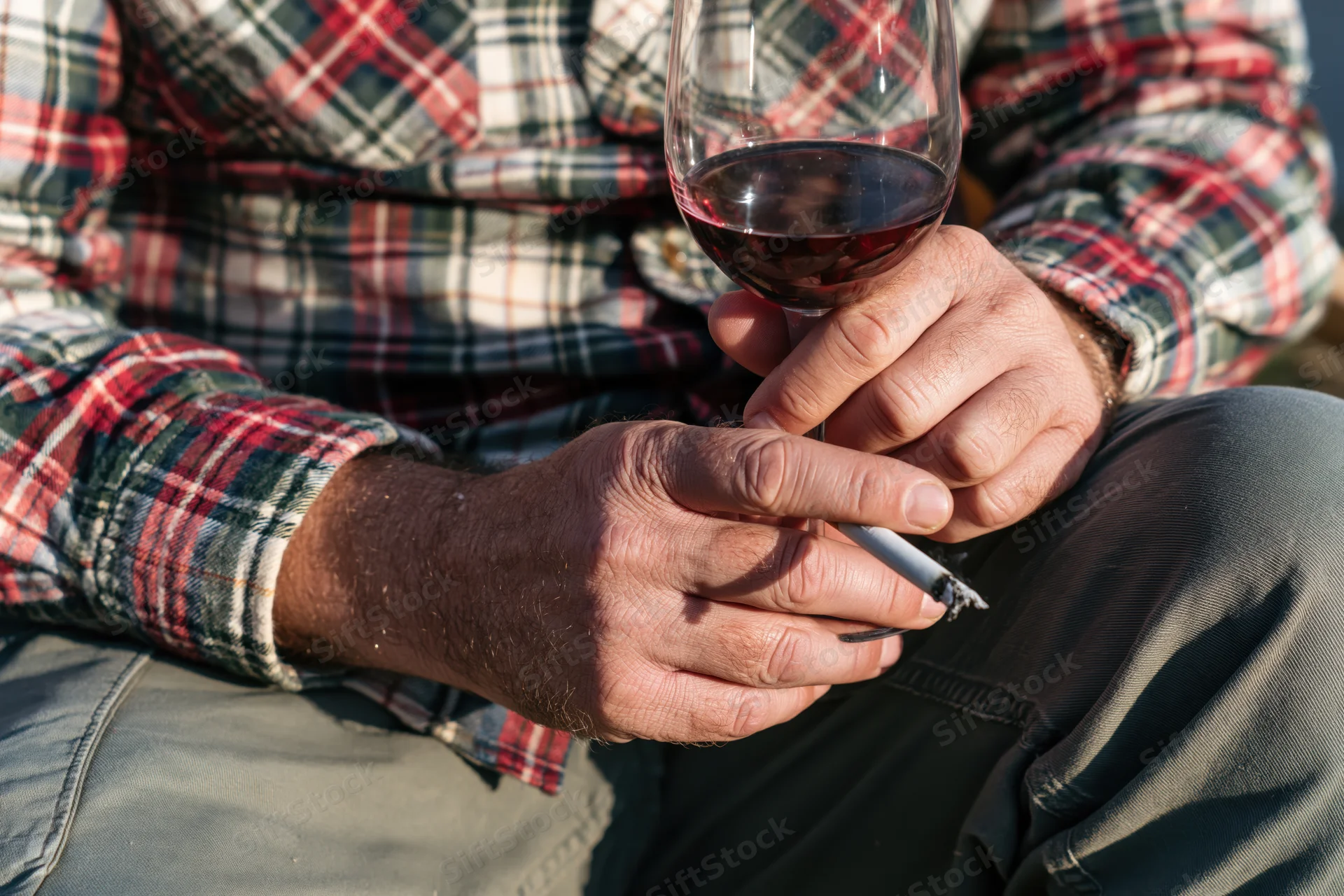 Man holding a glass of red wine and a cigarette while seated outdoors in a plaid shirt