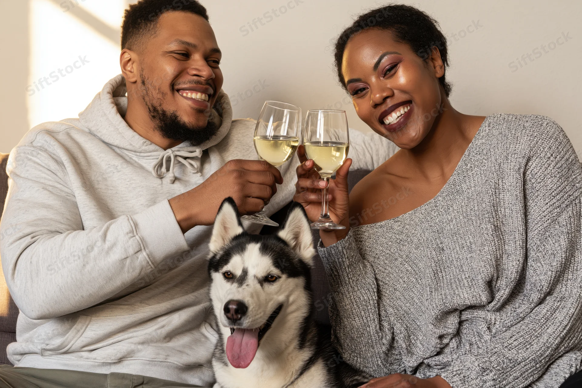 Couple toasting with wine glasses while sitting on couch with husky dog between them