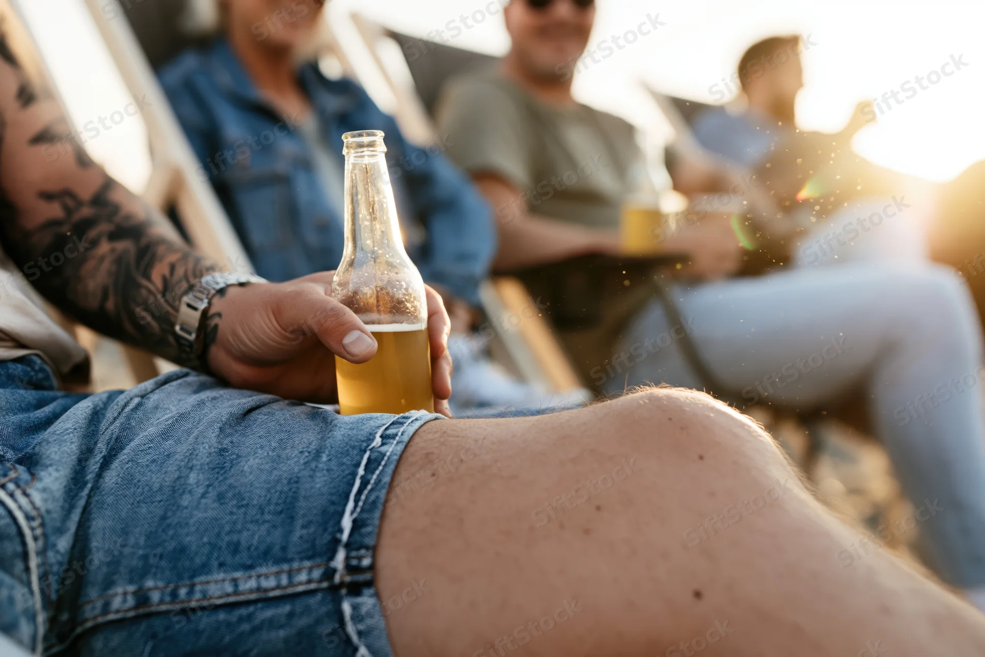 Man holding a beer bottle while relaxing on a deckchair with friends at sunset