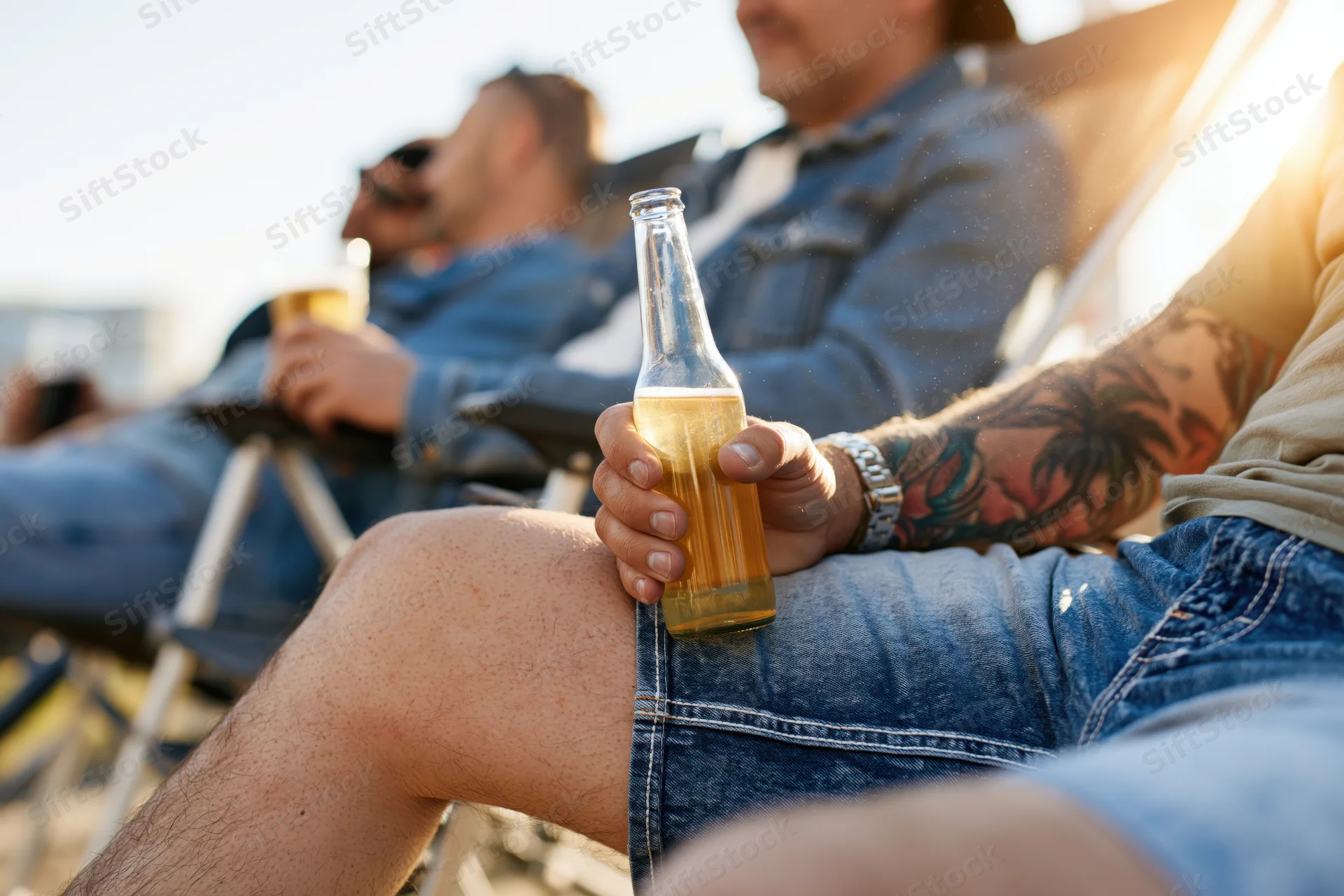 Man holding a beer bottle while relaxing with friends on a beach at sunset