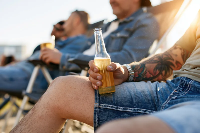 Man holding a beer bottle while relaxing with friends on a beach at sunset