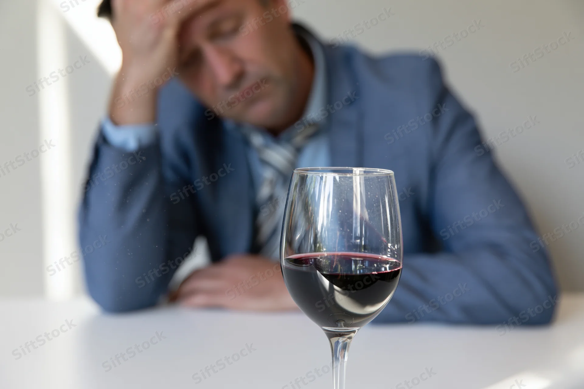 Man in suit resting head on hand beside a glass of red wine on a white table