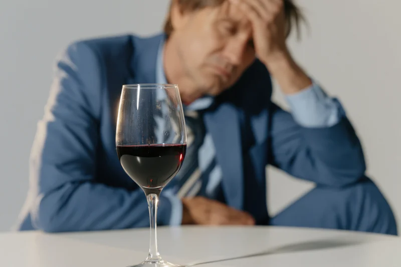 Wine glass with red wine in focus on white table, man in blue suit holding head in background